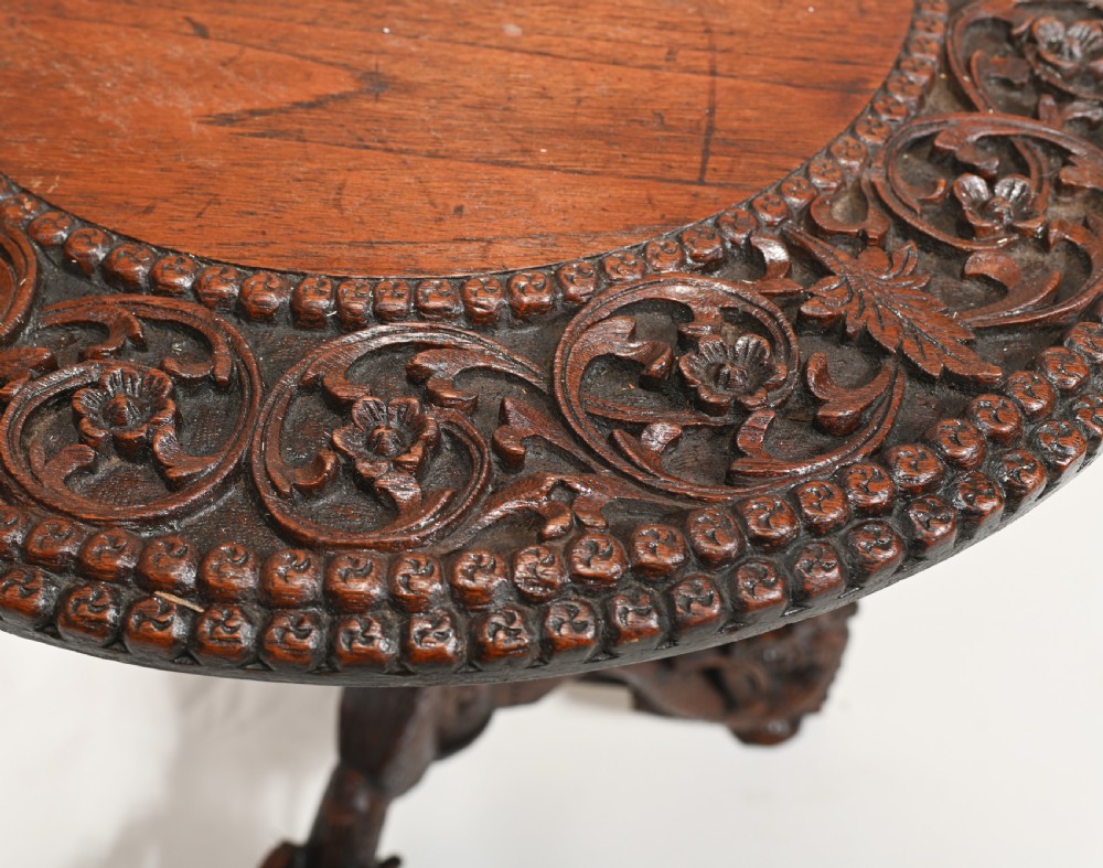 A dark brown carved timber side table featuring a circular top with a pierced floral border, supported on a turned stem and tripod legs shaped like mythological beasts. View VIII