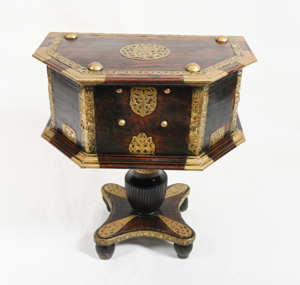 An antique dark wood Indian dowry casket decorated with intricate pierced brass straps and studs, supported on a turned central pedestal base.