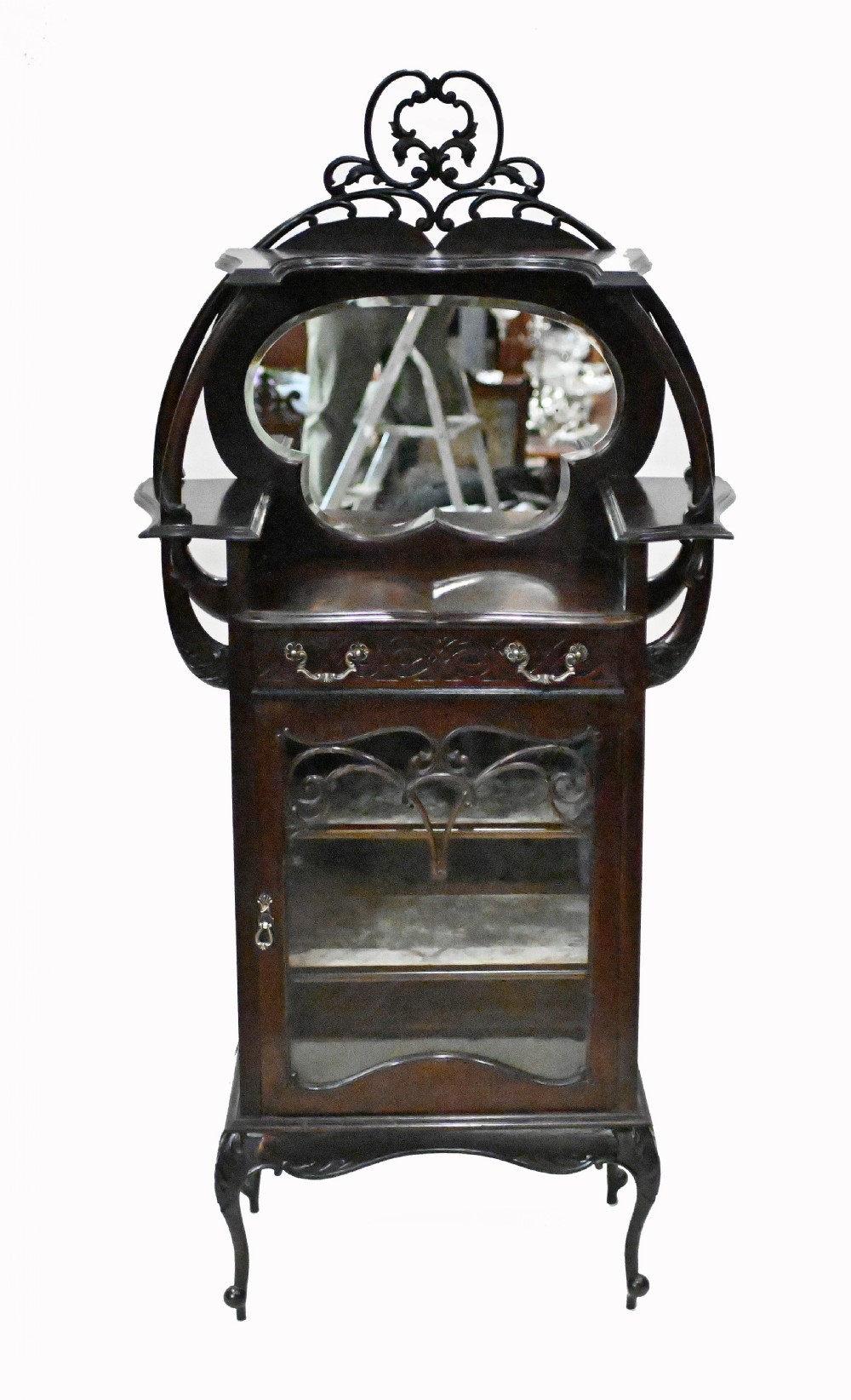 A dark mahogany Art Nouveau display cabinet featuring a pierced cresting, an asymmetrical mirrored superstructure, a single drawer, and a glazed door on slender shaped legs.