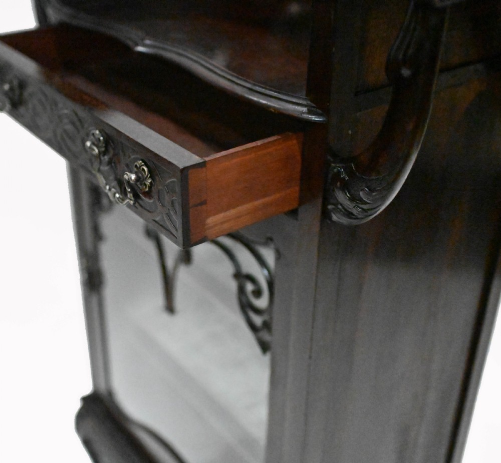A dark mahogany Art Nouveau display cabinet featuring a pierced cresting, an asymmetrical mirrored superstructure, a single drawer, and a glazed door on slender shaped legs. View VI