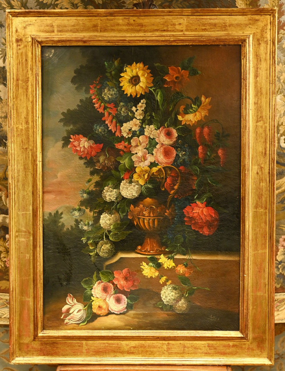 An antique oil painting depicting an elaborate arrangement of colourful flowers in a classical urn, set within a wide gilded wooden frame.