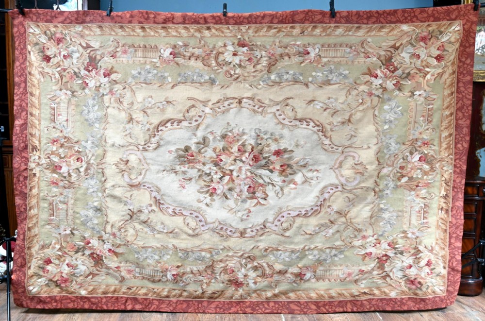 A large rectangular French Aubusson style flat-weave tapestry featuring a central floral bouquet medallion surrounded by scrolling foliate borders in pink, beige, and muted green shades.