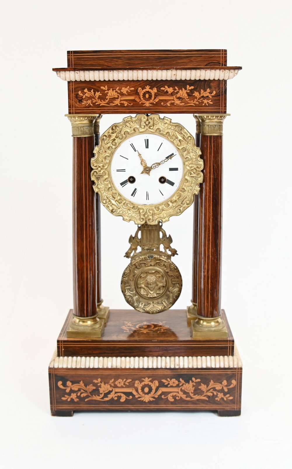 A French Charles X portico clock crafted from dark brown inlaid wood with four fluted columns, gilt bronze accents, a white enamel dial with Roman numerals, and a hanging brass pendulum.
