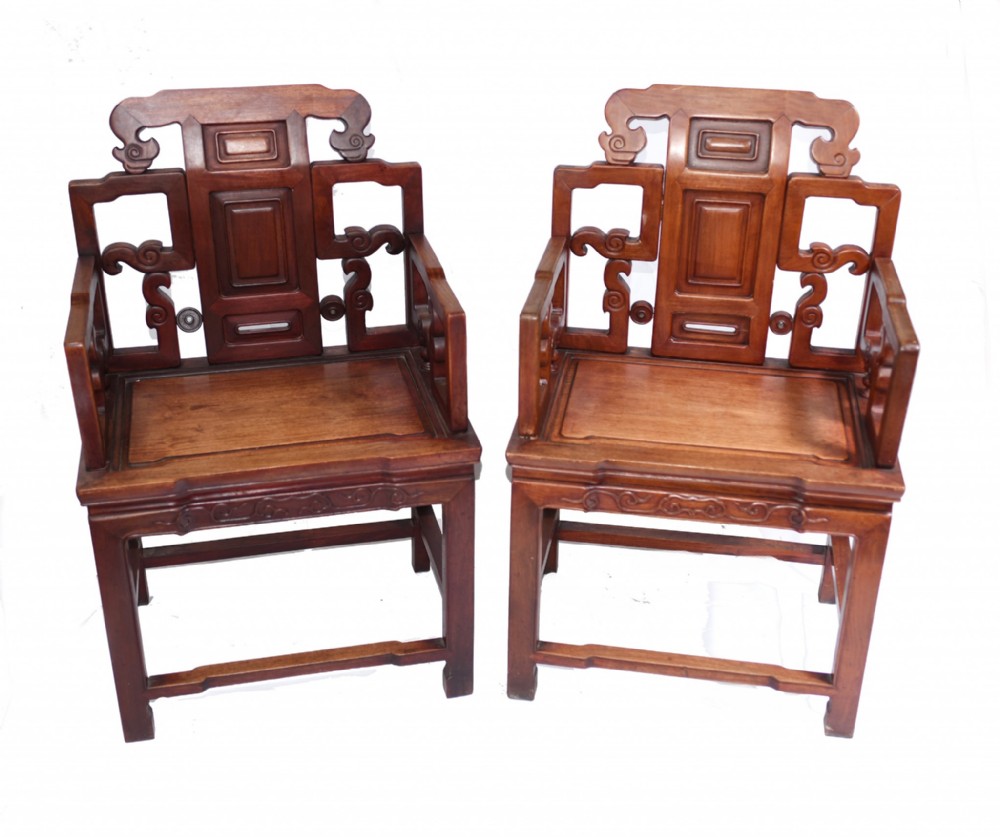 A matching pair of dark warm brown Chinese carved hardwood armchairs featuring decorative splats, openwork armrests, and hard wooden seats.