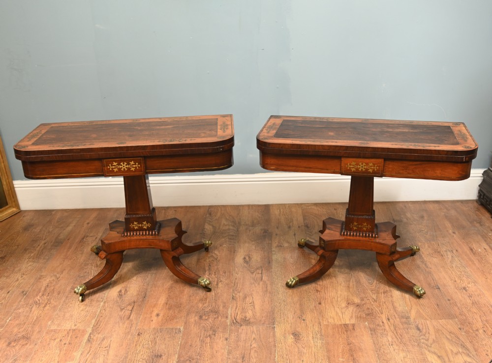 A pair of Regency rosewood and mahogany fold-over card tables featuring brass foliate inlay, central pedestal stems, and splayed quadform legs with brass paw castors.