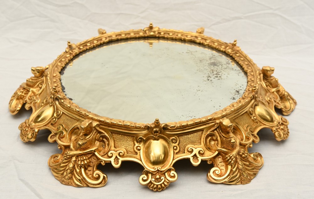 A circular French Rococo style gilt bronze mirrored plateau featuring an ornate scrollwork frame with decorative feet and a foxed mirror insert.