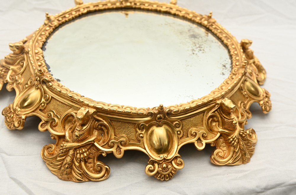 A circular French Rococo style gilt bronze mirrored plateau featuring an ornate scrollwork frame with decorative feet and a foxed mirror insert. View II