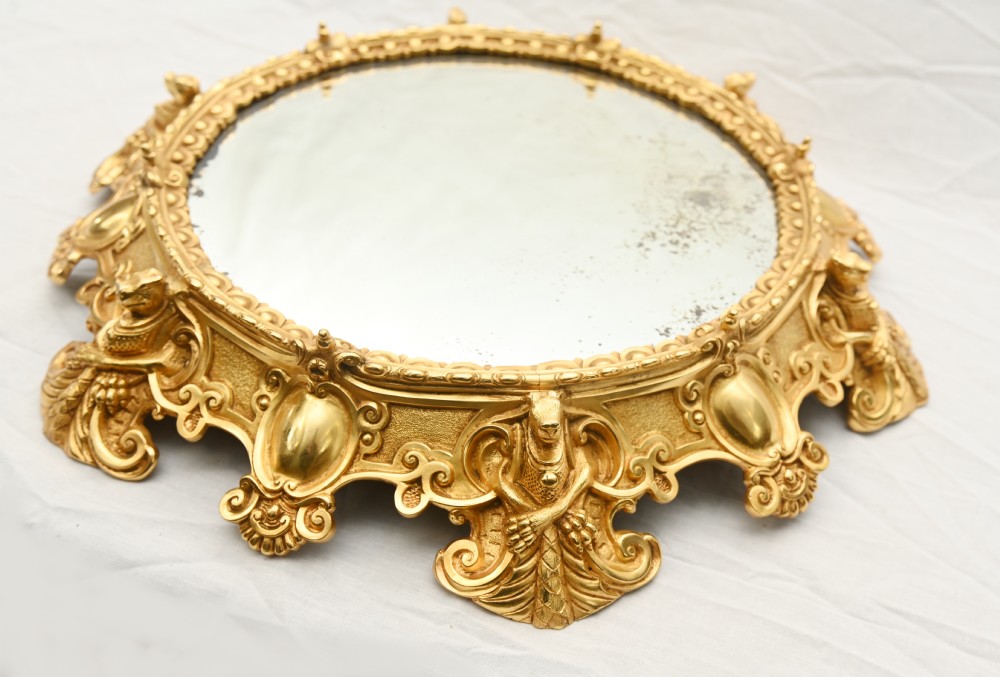 A circular French Rococo style gilt bronze mirrored plateau featuring an ornate scrollwork frame with decorative feet and a foxed mirror insert. View III