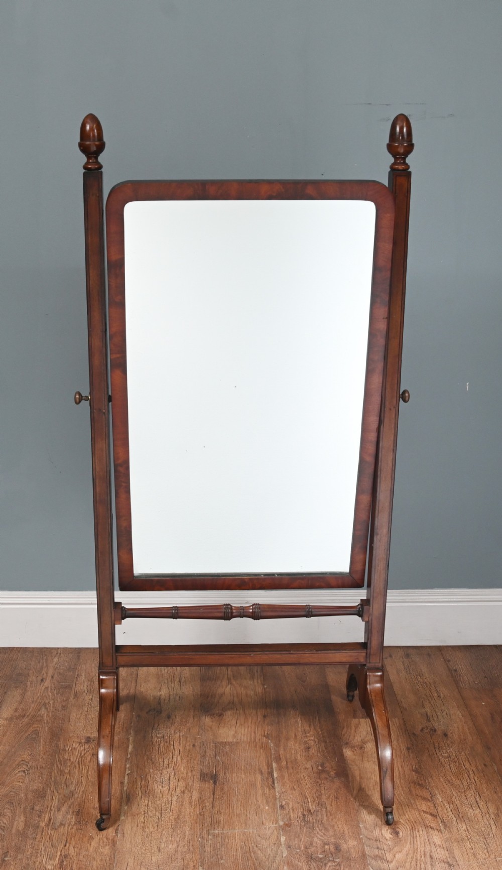 A Regency mahogany cheval mirror featuring an adjustable rectangular mirror plate, turned urn finials, a turned stretcher, and splayed legs on brass castors.
