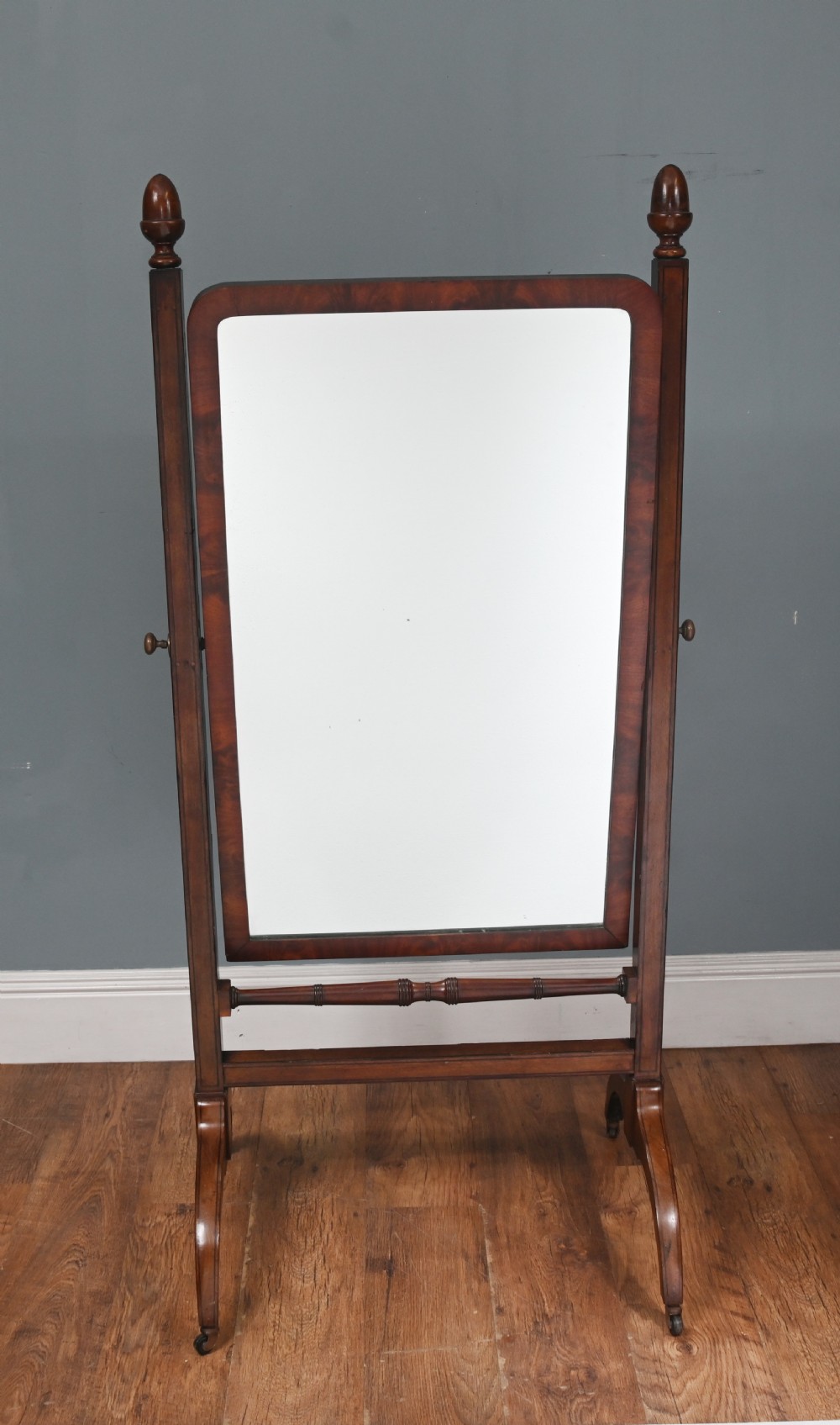 A Regency mahogany cheval mirror featuring an adjustable rectangular mirror plate, turned urn finials, a turned stretcher, and splayed legs on brass castors. View II