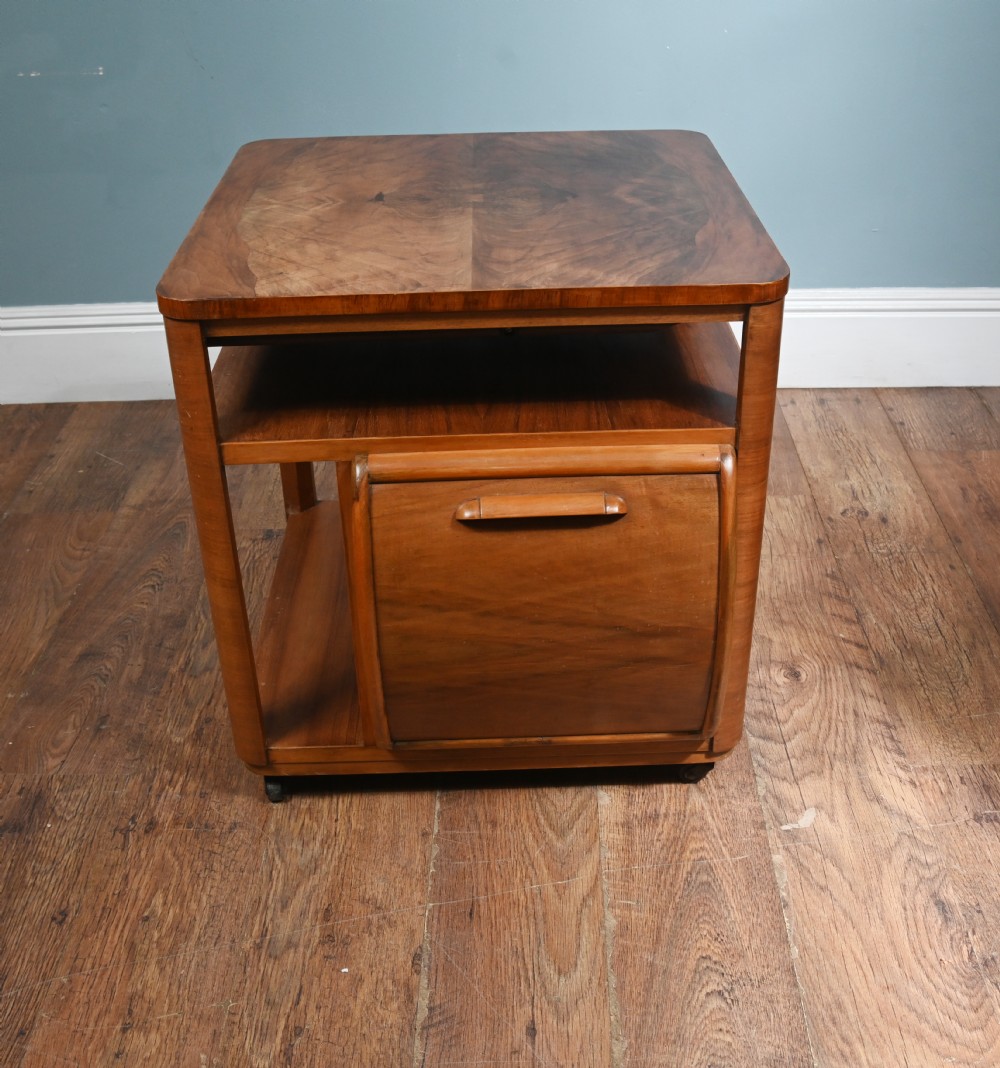 An Art Deco walnut side table featuring a square top, open shelving, a central drop-front cabinet compartment, and small base castors.