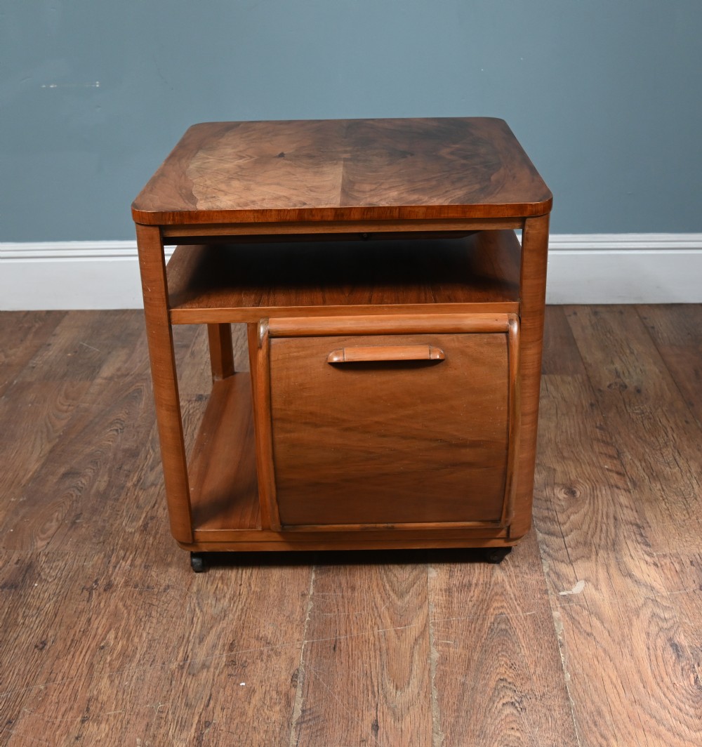 An Art Deco walnut side table featuring a square top, open shelving, a central drop-front cabinet compartment, and small base castors. View II