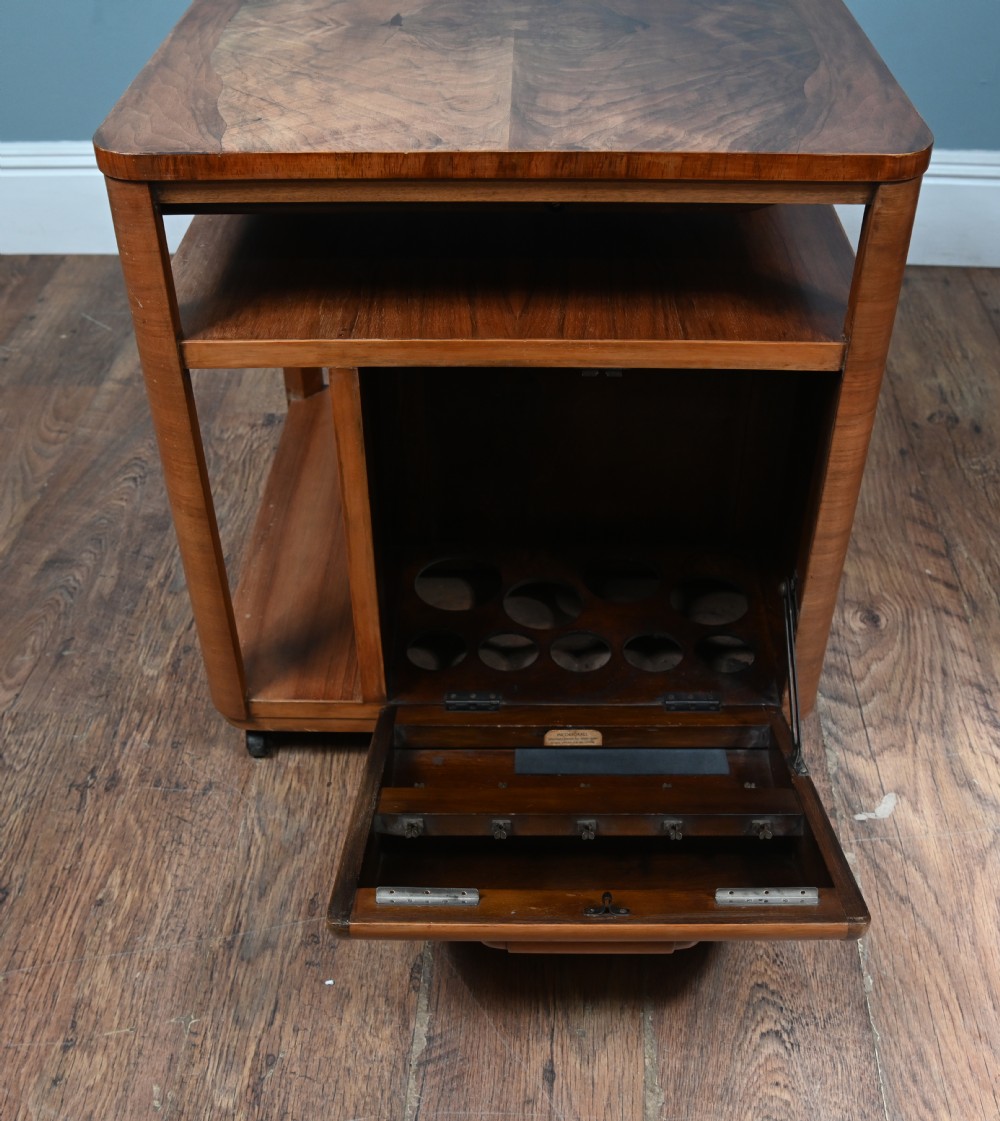 An Art Deco walnut side table featuring a square top, open shelving, a central drop-front cabinet compartment, and small base castors. View IV