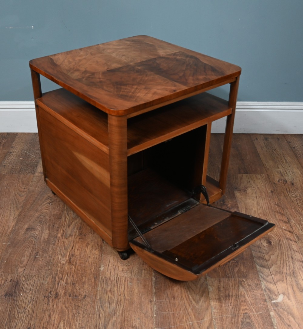An Art Deco walnut side table featuring a square top, open shelving, a central drop-front cabinet compartment, and small base castors. View VIII