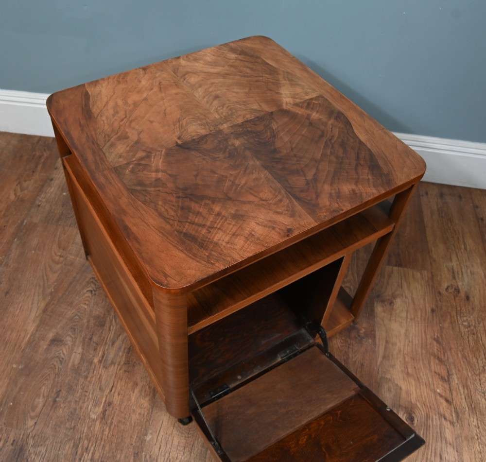An Art Deco walnut side table featuring a square top, open shelving, a central drop-front cabinet compartment, and small base castors. View IX