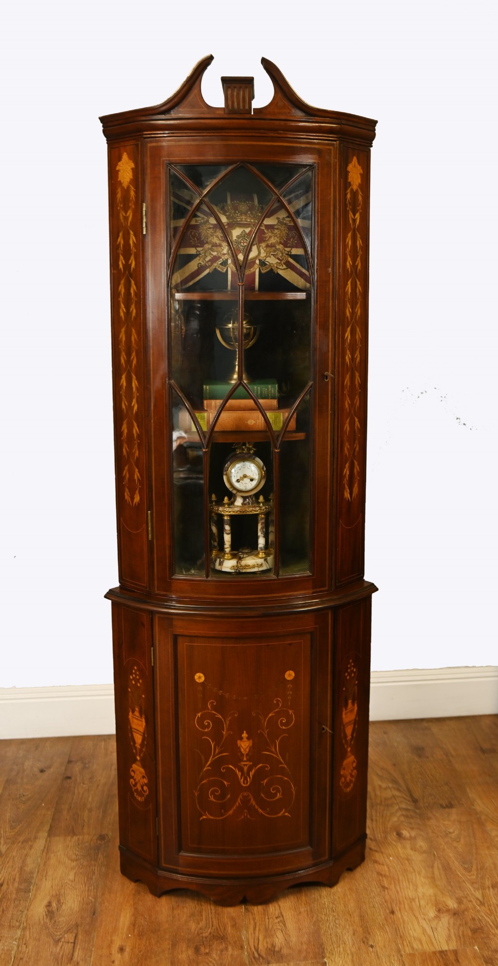 An Edwardian inlaid mahogany corner display cabinet featuring a swan-neck pediment, astragal glazed upper door, and marquetry urn designs on the lower panel.