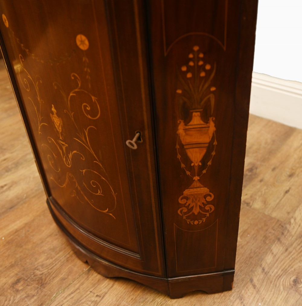 An Edwardian inlaid mahogany corner display cabinet featuring a swan-neck pediment, astragal glazed upper door, and marquetry urn designs on the lower panel. View VI