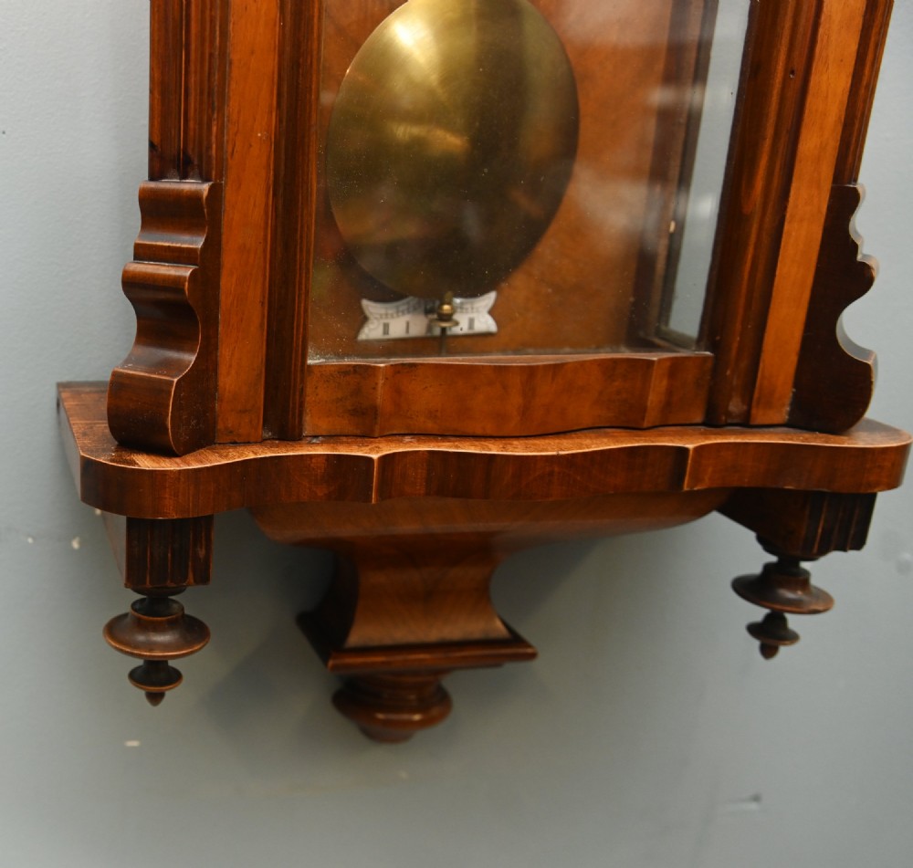 An antique walnut Vienna regulator wall clock featuring a glazed front door, brass pendulum, and turned bottom finials. View V