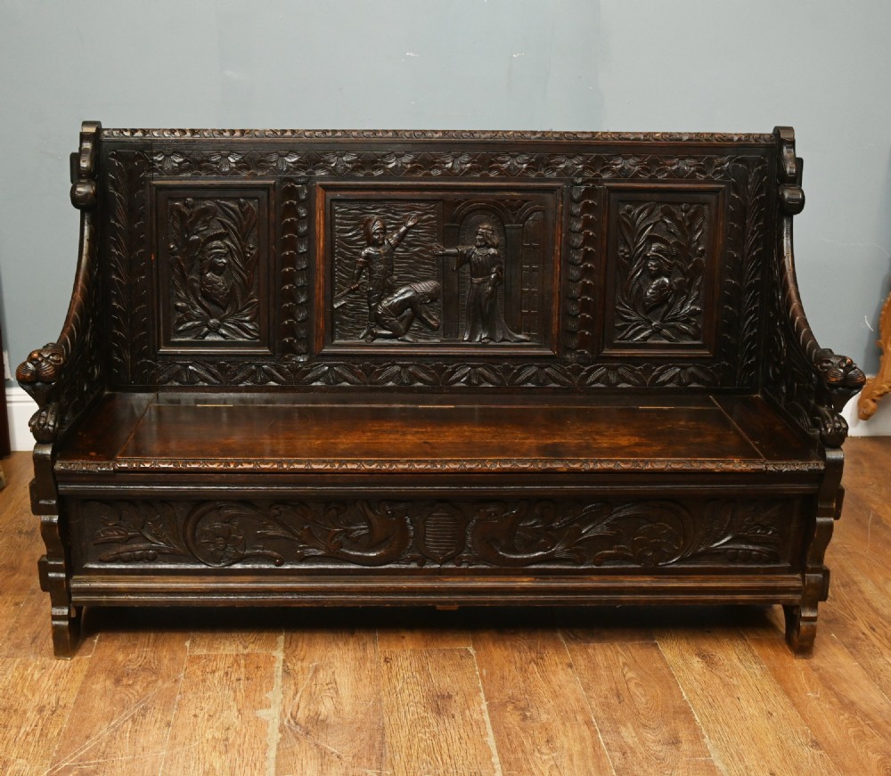 A dark Renaissance Revival carved oak hall settle featuring narrative relief panels on the backrest, carved lion head armrests, and a lifted storage seat. View II