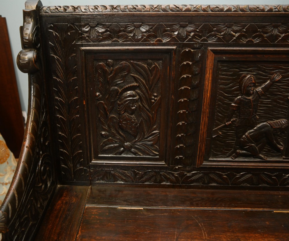 A dark Renaissance Revival carved oak hall settle featuring narrative relief panels on the backrest, carved lion head armrests, and a lifted storage seat. View IV