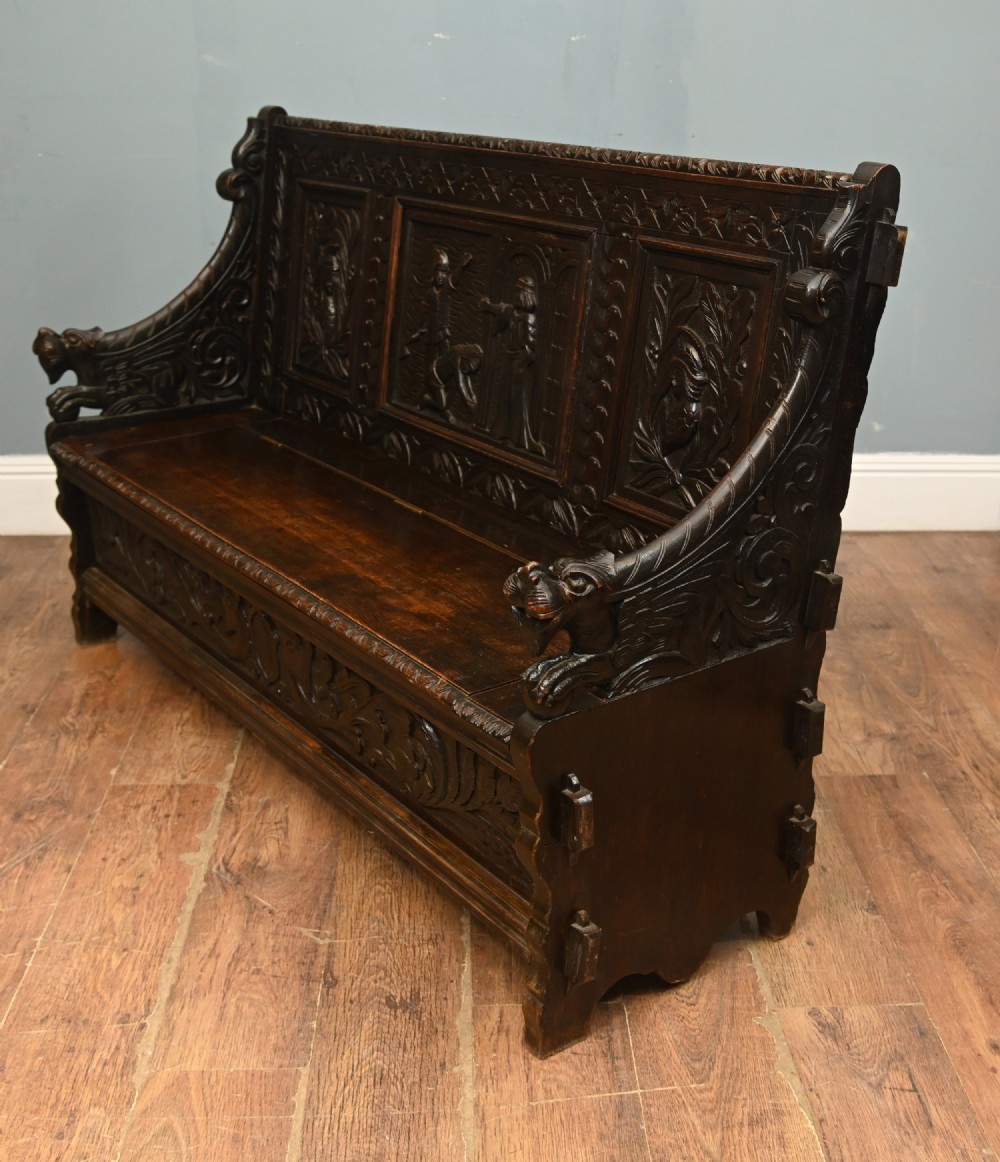 A dark Renaissance Revival carved oak hall settle featuring narrative relief panels on the backrest, carved lion head armrests, and a lifted storage seat. View V