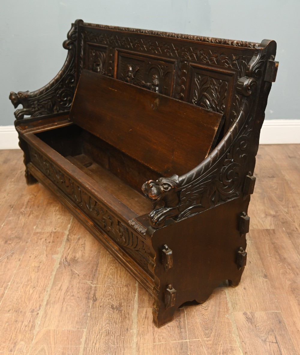 A dark Renaissance Revival carved oak hall settle featuring narrative relief panels on the backrest, carved lion head armrests, and a lifted storage seat. View VI