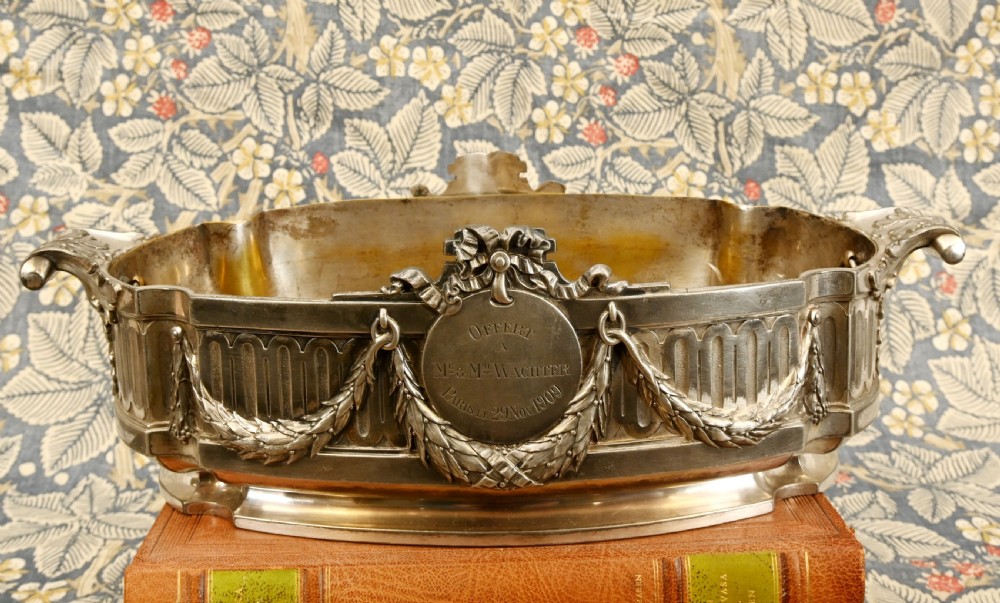 An oval Neoclassical silver-plated jardiniere featuring ribbon bow motifs, hanging laurel swags, twin end handles, and an engraved central medallion dated 1909.