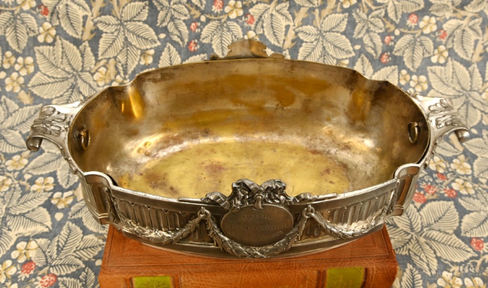 An oval Neoclassical silver-plated jardiniere featuring ribbon bow motifs, hanging laurel swags, twin end handles, and an engraved central medallion dated 1909. View IV