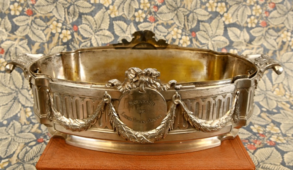 An oval Neoclassical silver-plated jardiniere featuring ribbon bow motifs, hanging laurel swags, twin end handles, and an engraved central medallion dated 1909. View VI