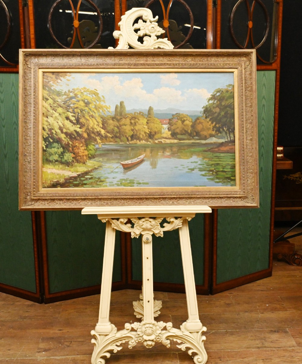 A framed French impressionist oil painting depicting a river scene with a wooden boat moored near tree-lined banks under a blue sky.