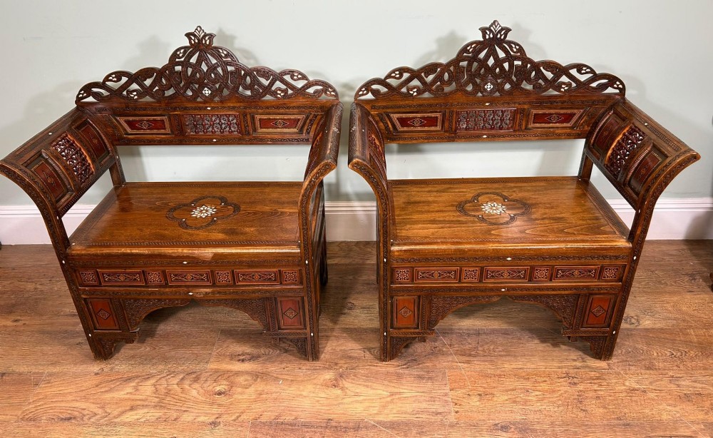 A pair of warm brown wooden armchairs featuring elaborate carved openwork crestings and geometric mother-of-pearl inlays across the seats and panels.