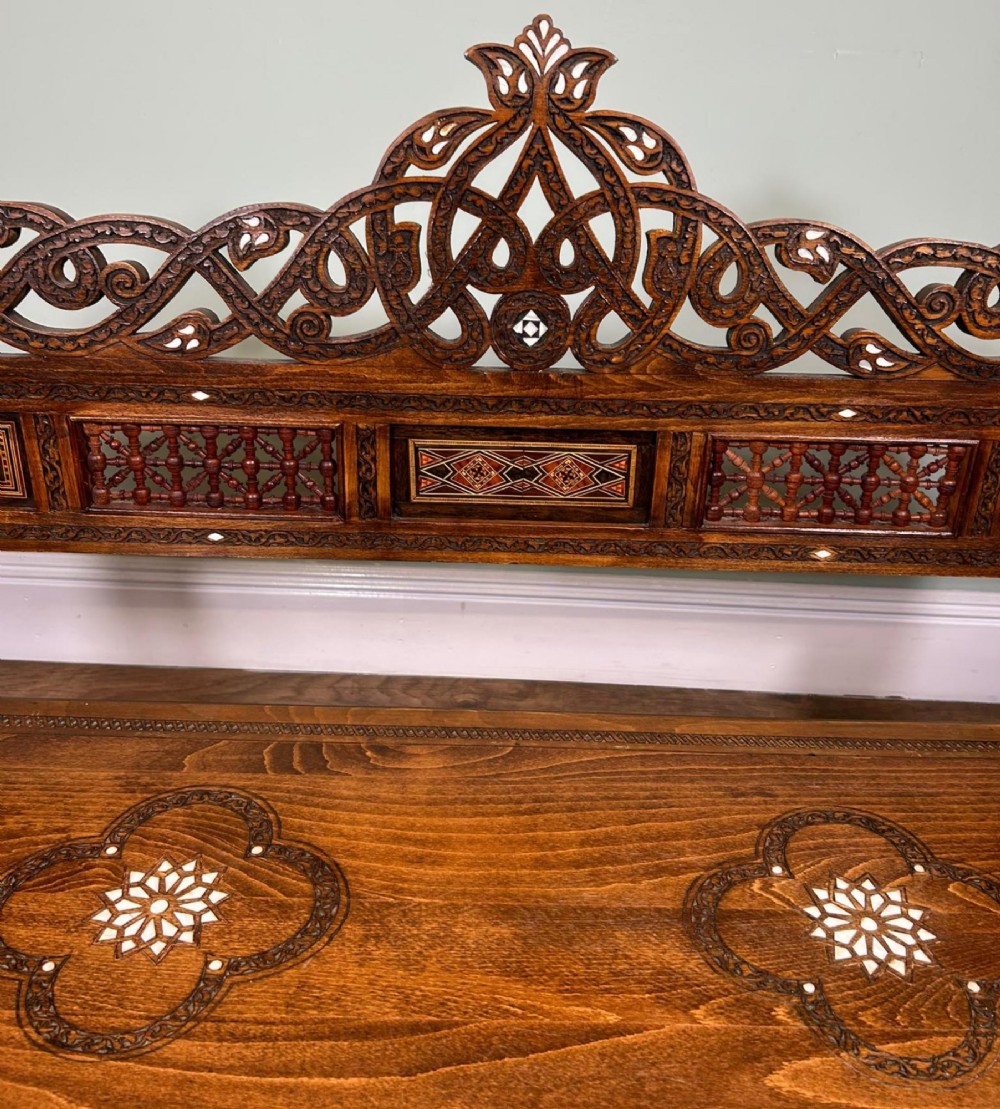 A 19th-century Syrian wooden bench featuring geometric marquetry inlay, mother-of-pearl accents, an openwork carved crest, and arched apron support. View IV