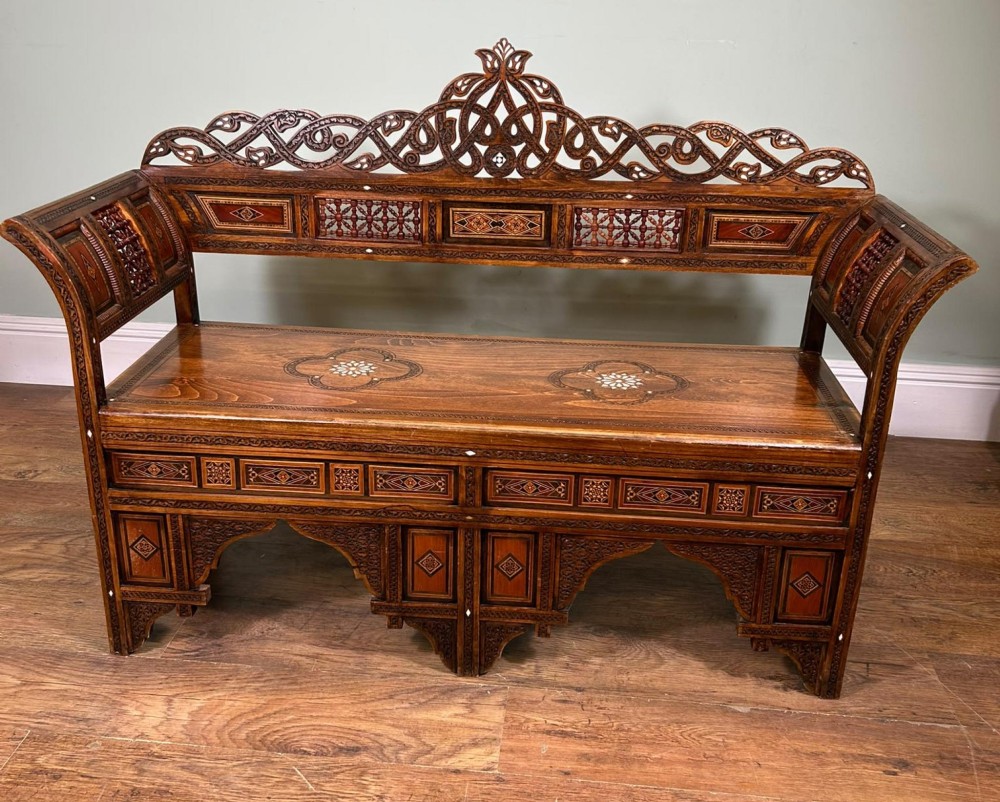 A 19th-century Syrian wooden bench featuring geometric marquetry inlay, mother-of-pearl accents, an openwork carved crest, and arched apron support. View V