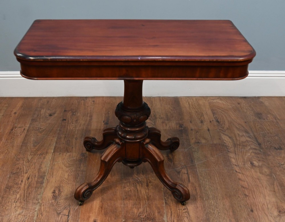A Victorian mahogany fold-over card table with a rectangular top supported by a central carved pedestal and four scroll-cut legs on castors.