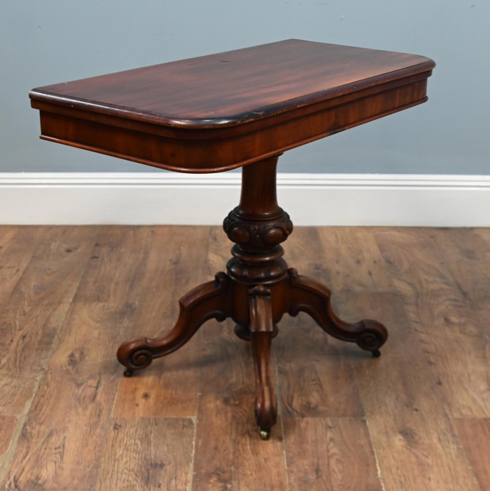 A Victorian mahogany fold-over card table with a rectangular top supported by a central carved pedestal and four scroll-cut legs on castors. View II