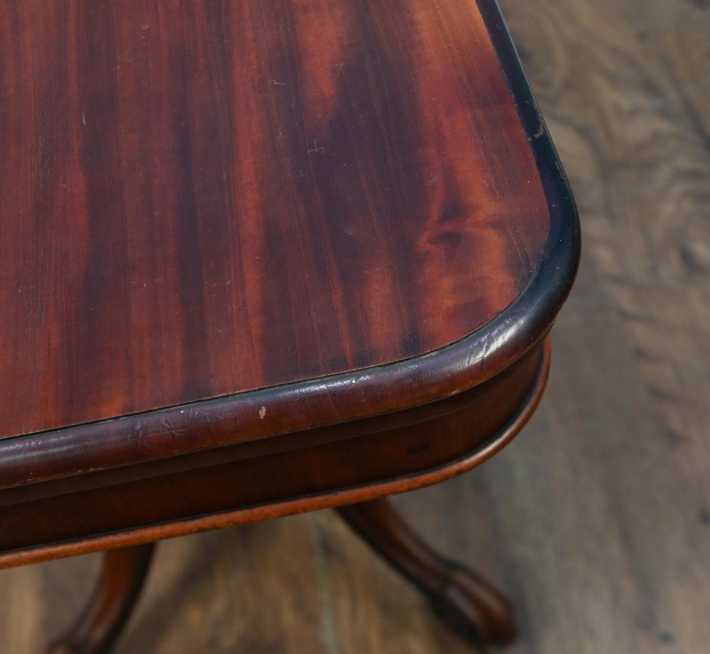 A Victorian mahogany fold-over card table with a rectangular top supported by a central carved pedestal and four scroll-cut legs on castors. View V