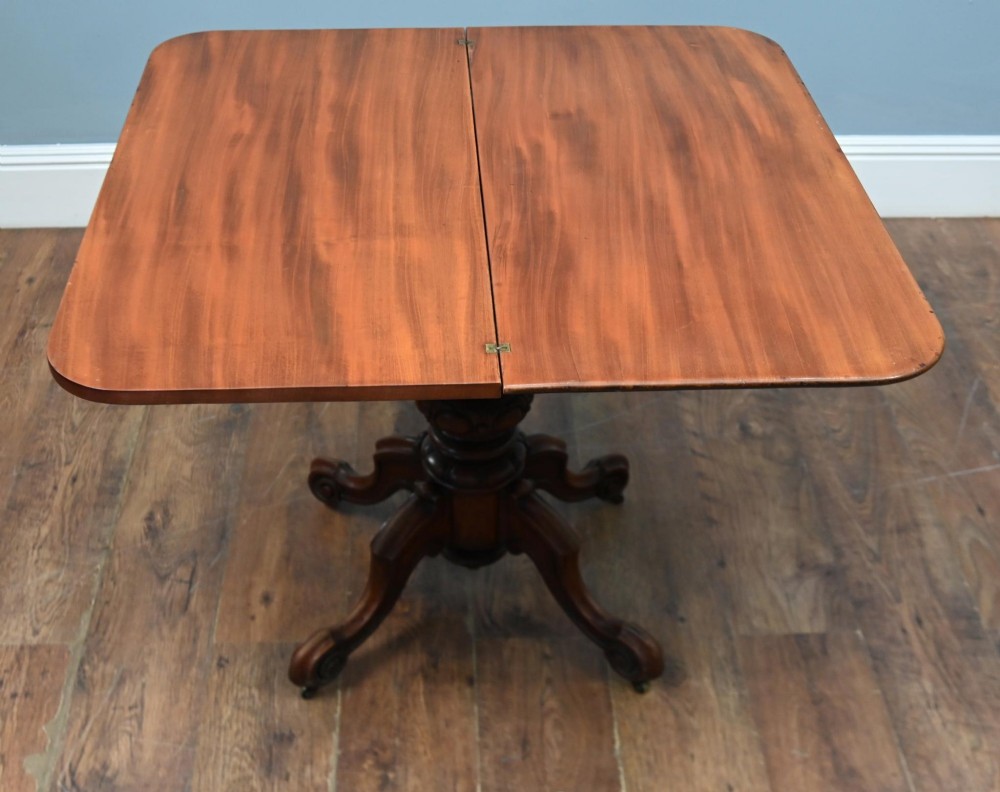 A Victorian mahogany fold-over card table with a rectangular top supported by a central carved pedestal and four scroll-cut legs on castors. View IX