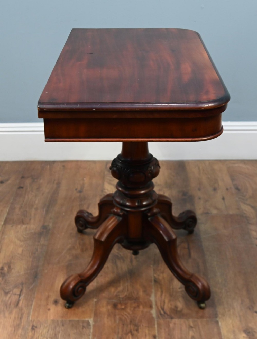 A Victorian mahogany fold-over card table with a rectangular top supported by a central carved pedestal and four scroll-cut legs on castors. View XI