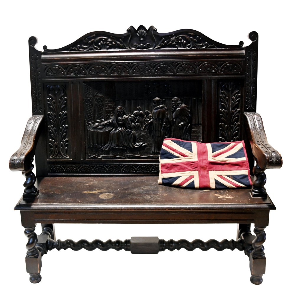 A late 19th-century dark carved walnut hall bench featuring a pictorial relief back panel, scrolled armrests, and turned stretchers.