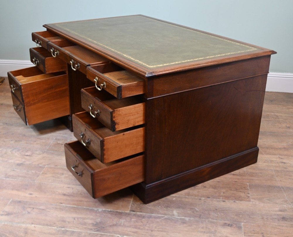 A Victorian mahogany pedestal desk featuring nine brass-handled drawers and a green leather gilt-tooled writing surface resting on a solid plinth base. View IX