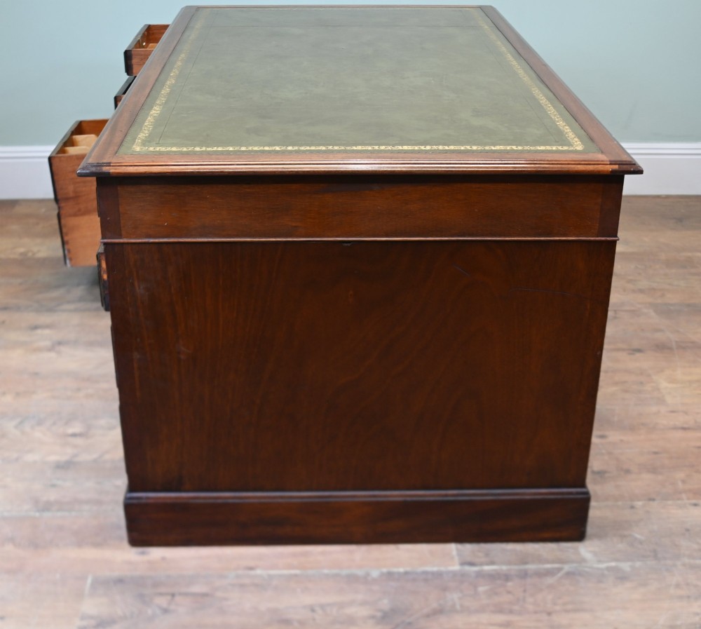 A Victorian mahogany pedestal desk featuring nine brass-handled drawers and a green leather gilt-tooled writing surface resting on a solid plinth base. View X