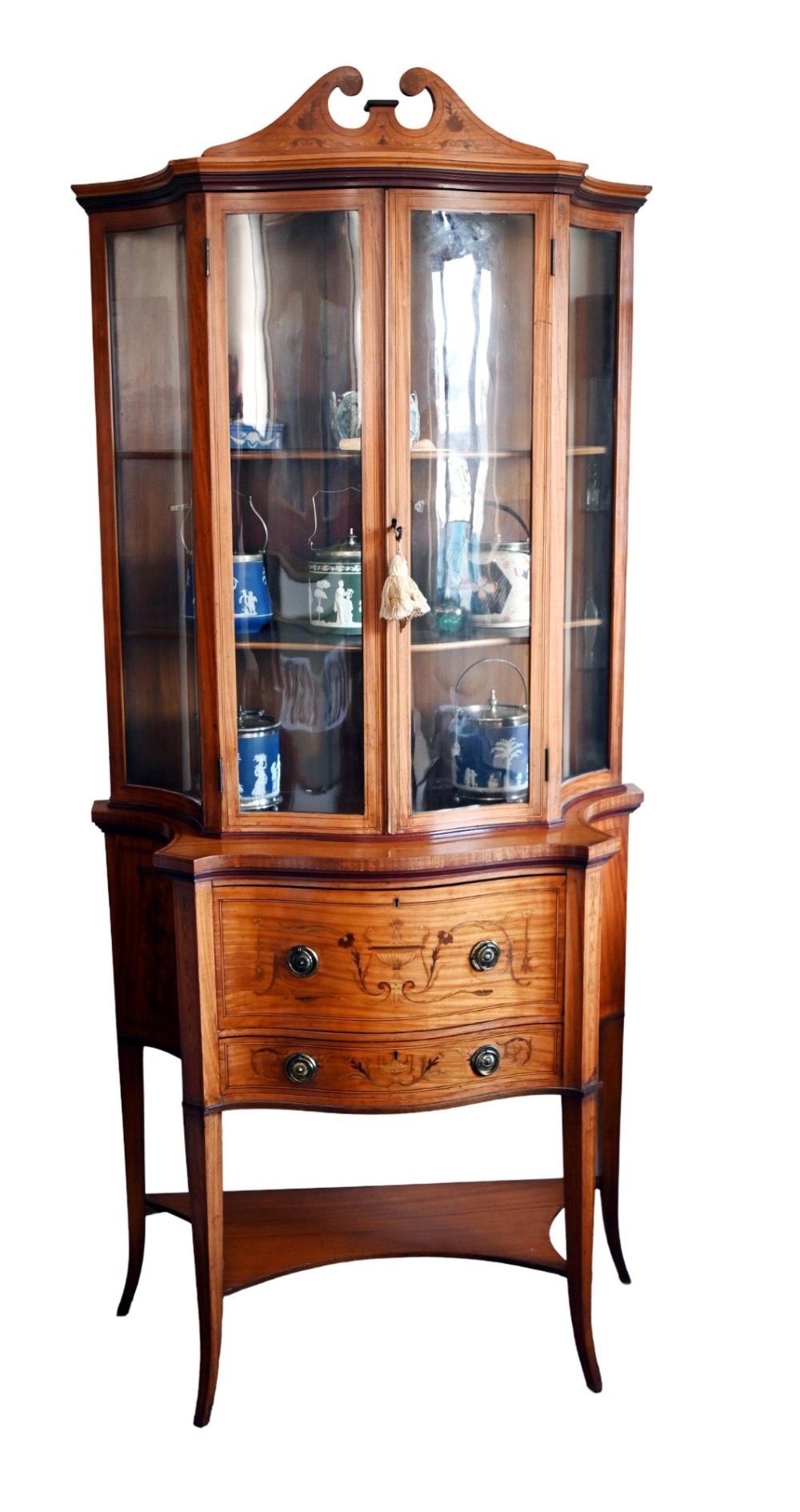 An Edwardian satinwood glazed display cabinet featuring classical urn marquetry, a swan-neck pediment, bowed glass sides, and drawers over tapered legs.