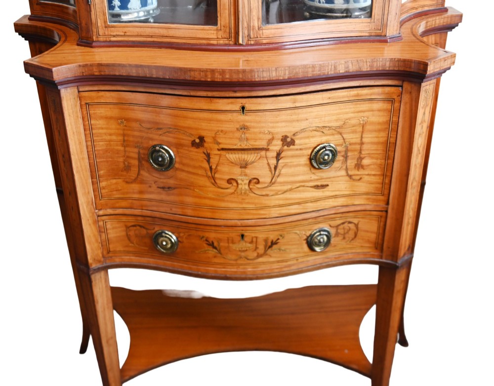 An Edwardian satinwood glazed display cabinet featuring classical urn marquetry, a swan-neck pediment, bowed glass sides, and drawers over tapered legs. View II