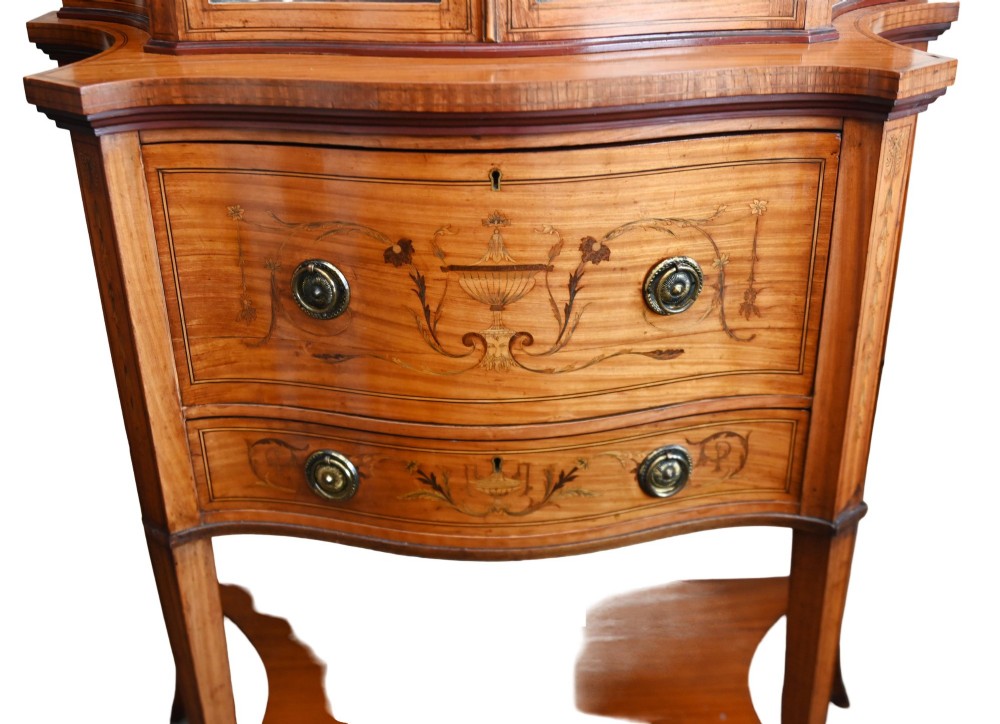 An Edwardian satinwood glazed display cabinet featuring classical urn marquetry, a swan-neck pediment, bowed glass sides, and drawers over tapered legs. View IV