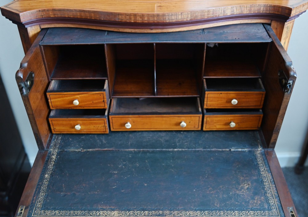 An Edwardian satinwood glazed display cabinet featuring classical urn marquetry, a swan-neck pediment, bowed glass sides, and drawers over tapered legs. View VIII