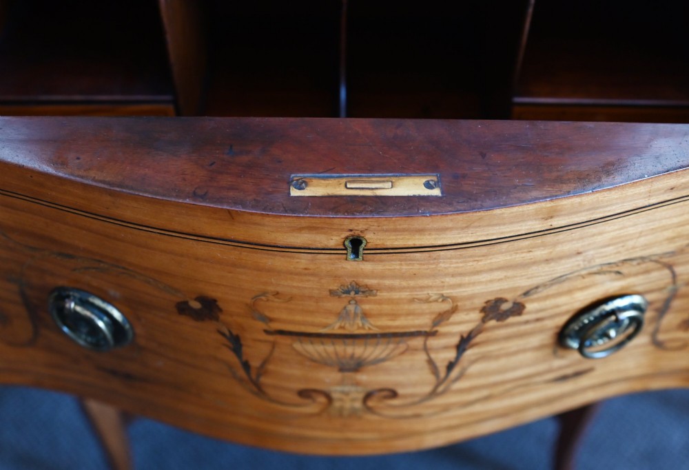 An Edwardian satinwood glazed display cabinet featuring classical urn marquetry, a swan-neck pediment, bowed glass sides, and drawers over tapered legs. View IX