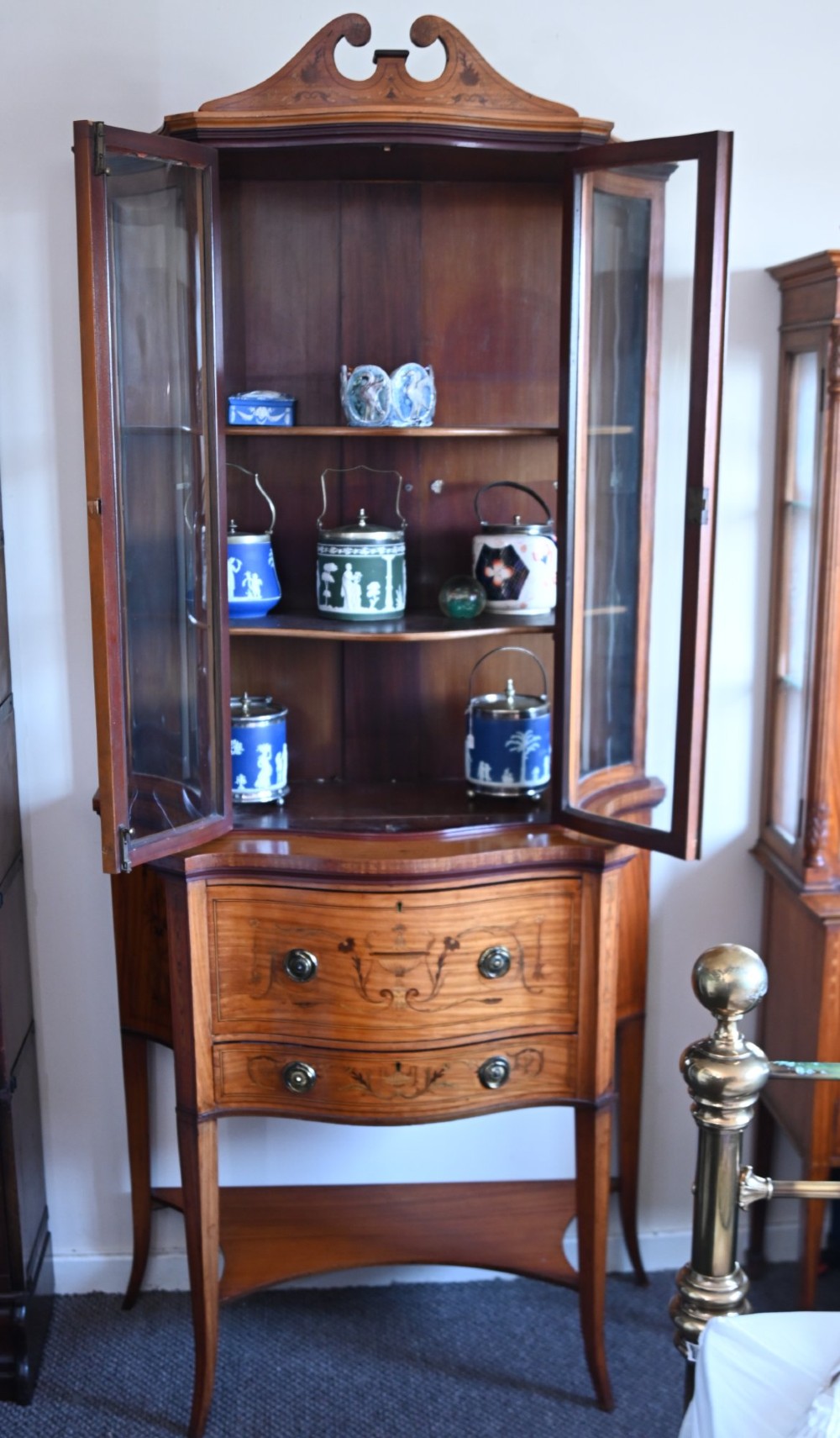 An Edwardian satinwood glazed display cabinet featuring classical urn marquetry, a swan-neck pediment, bowed glass sides, and drawers over tapered legs. View X