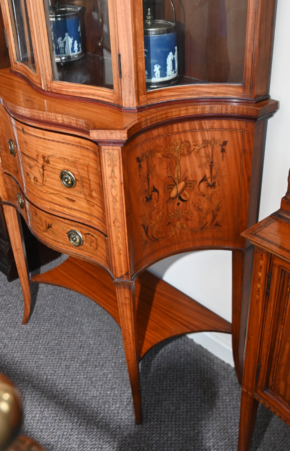 An Edwardian satinwood glazed display cabinet featuring classical urn marquetry, a swan-neck pediment, bowed glass sides, and drawers over tapered legs. View XII