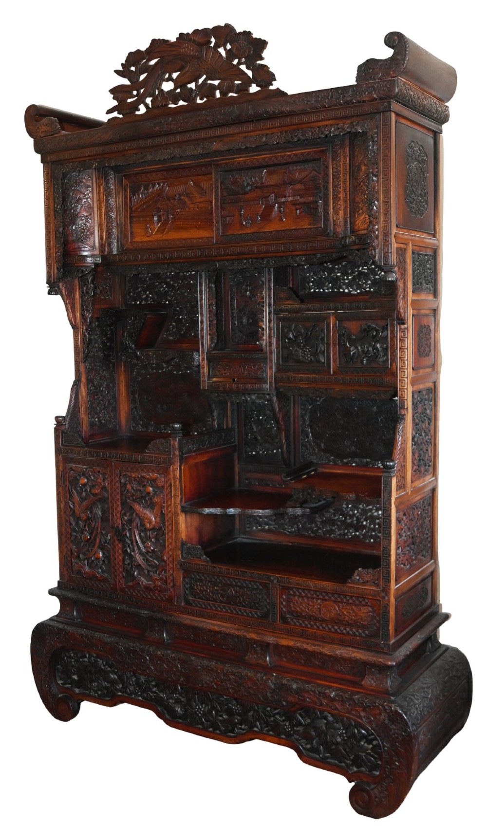 An intricately carved dark brown Japanese Meiji period display cabinet featuring asymmetrical open shelves, relief-carved cupboard doors, and a decorative cresting piece.