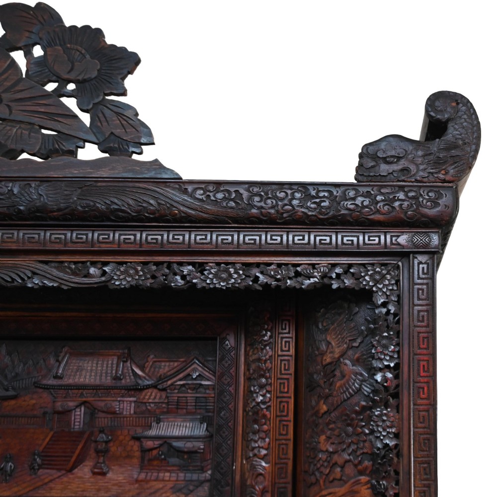 An intricately carved dark brown Japanese Meiji period display cabinet featuring asymmetrical open shelves, relief-carved cupboard doors, and a decorative cresting piece. View V
